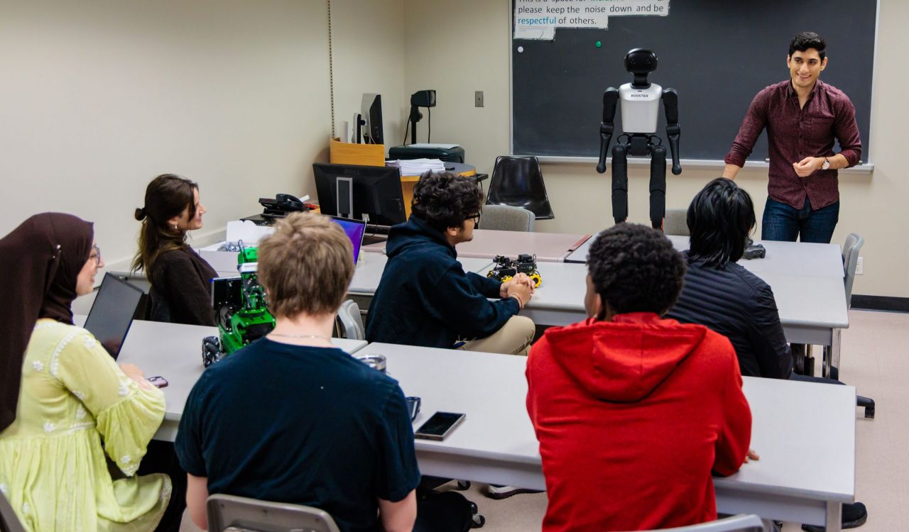 Students seated at tables engage in a hands-on robotics lesson while an instructor presents a humanoid robot, with computers and equipment supporting the learning environment.