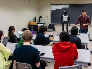 Students seated at tables engage in a hands-on robotics lesson while an instructor presents a humanoid robot, with computers and equipment supporting the learning environment.