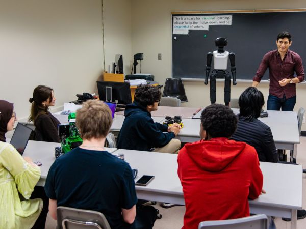 Students seated at tables engage in a hands-on robotics lesson while an instructor presents a humanoid robot, with computers and equipment supporting the learning environment.