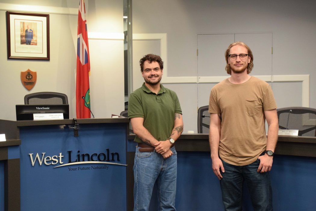 Two men stand beside each other in a municipal council chamber. 