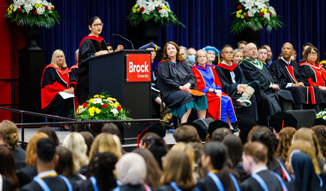 A woman stands at a podium speaking in a gymnasium filled with graduates.