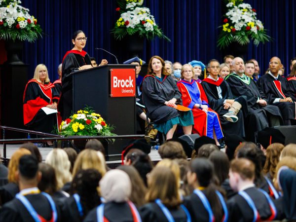 A woman stands at a podium speaking in a gymnasium filled with graduates.