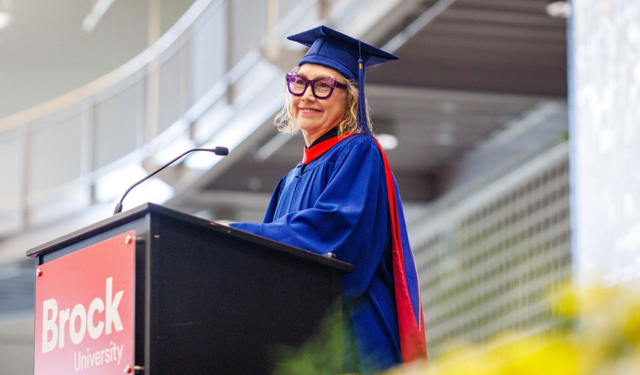 A woman in a Convocation gown stands at a podium smiling.
