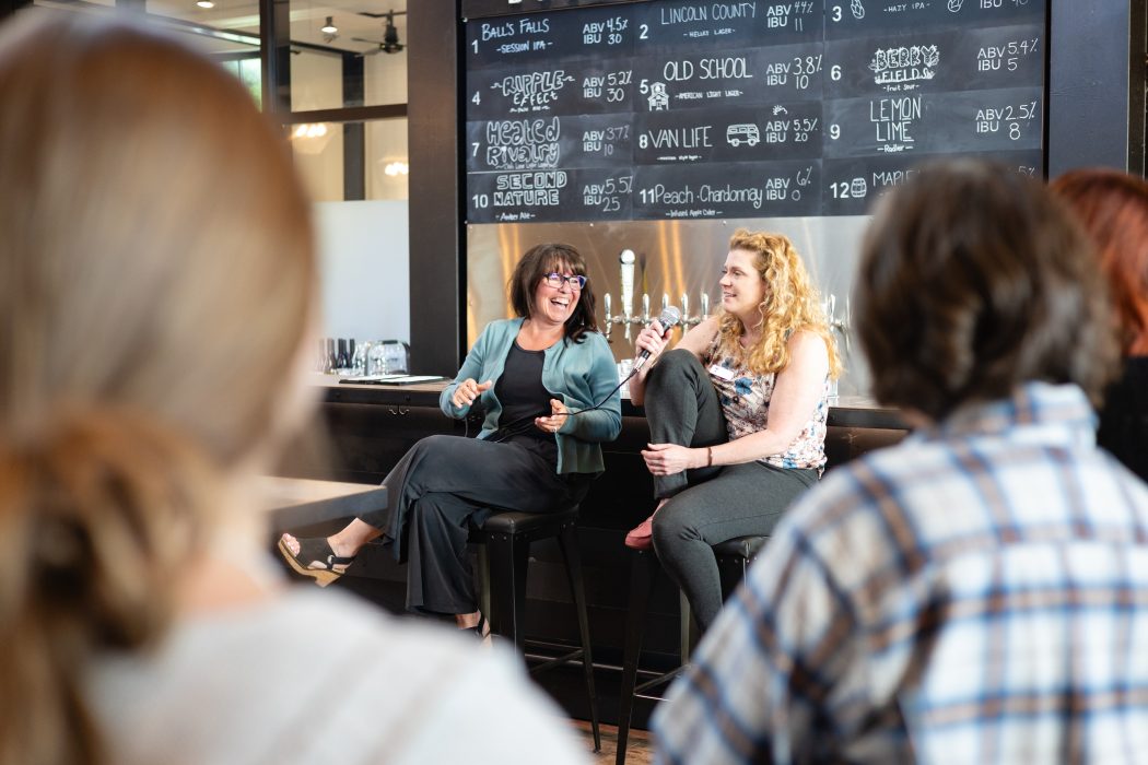 Two people holding mics and sitting on stools in front of a bar.