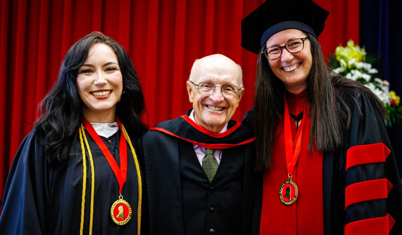Three smiling people in academic regalia pose together in front of a red curtain during a university convocation ceremony.