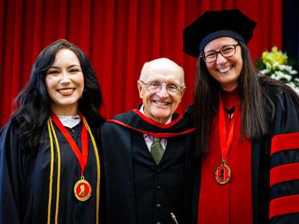 Three smiling people in academic regalia pose together in front of a red curtain during a university convocation ceremony.