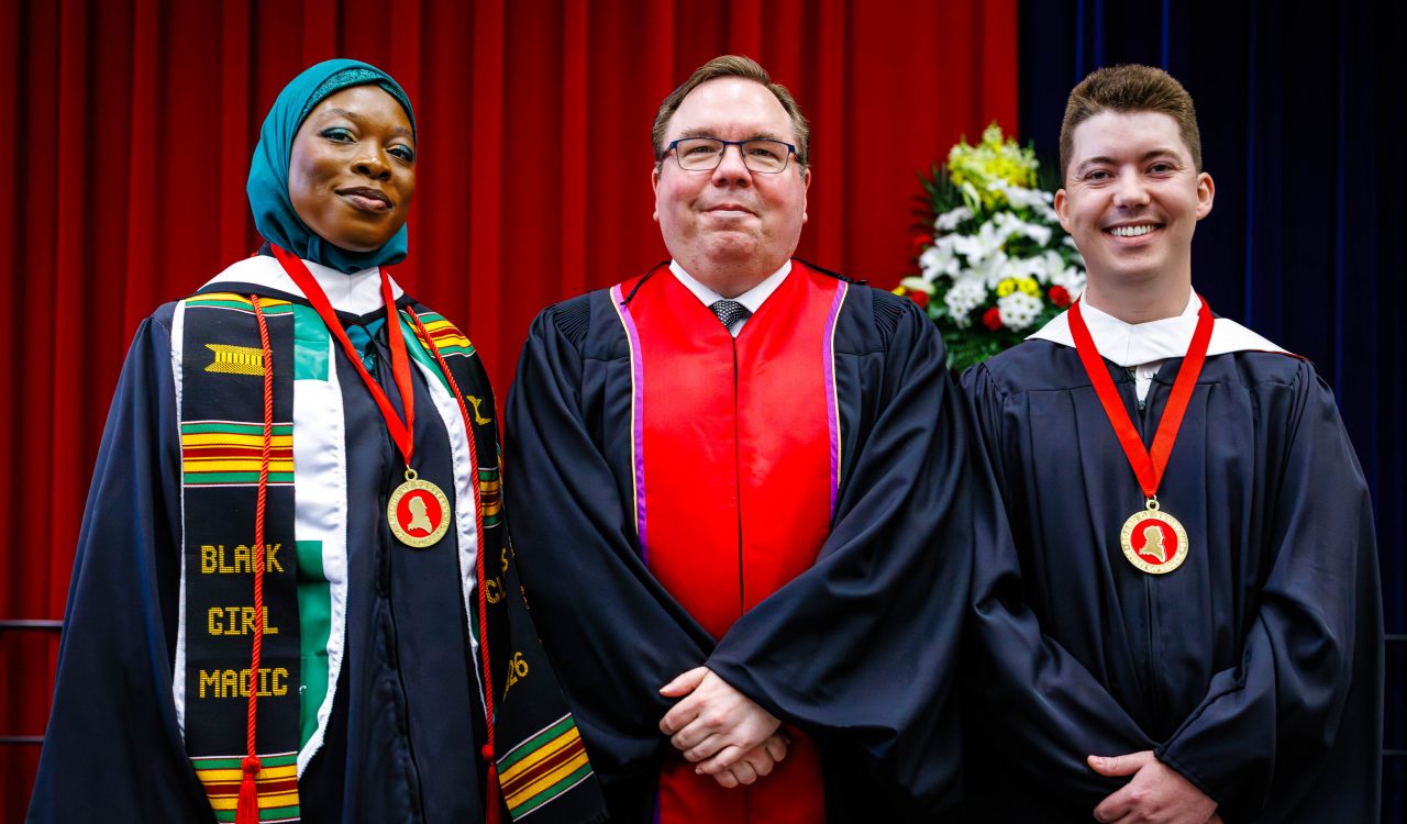 Three people in academic robes pose for a photo during a university graduation ceremony. Two of the people are wearing medals.