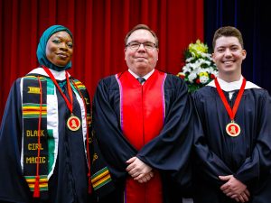 Three people in academic robes pose for a photo during a university graduation ceremony. Two of the people are wearing medals.