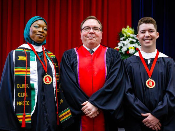 Three people in academic robes pose for a photo during a university graduation ceremony. Two of the people are wearing medals.