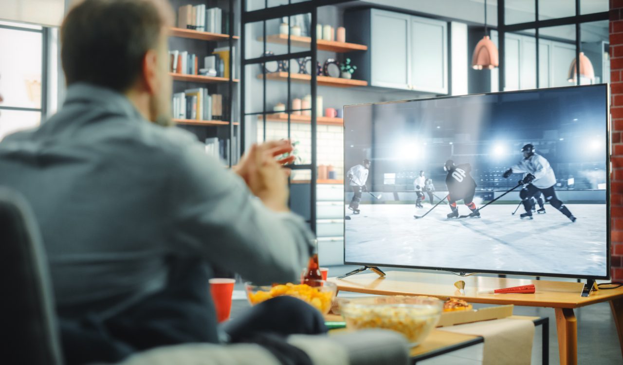 A man, seen from behind, watches ice hockey on a TV.