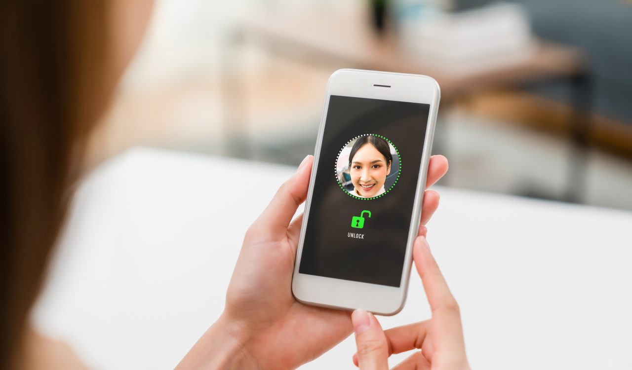 A woman holds a smartphone showing a photo of her face to unlock the device.