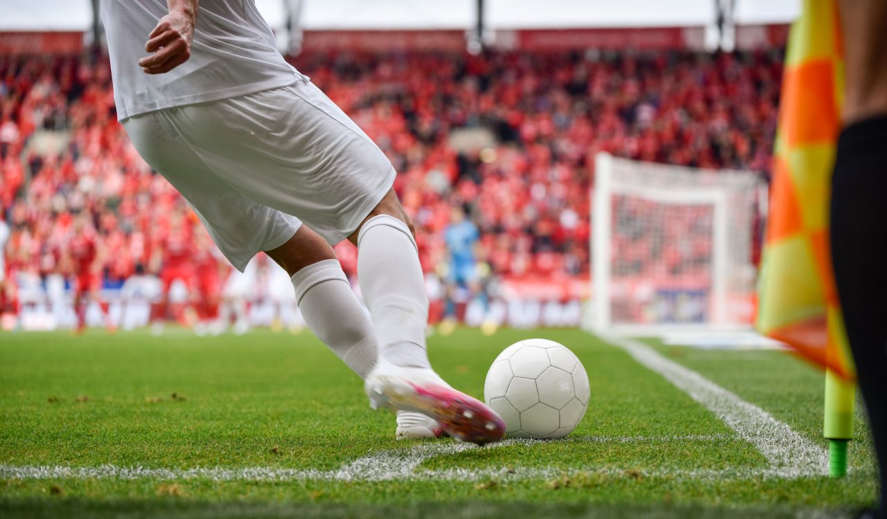 Close-up of the legs of a soccer player kicking a ball with a field and spectators in the background.