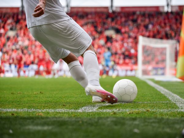 Close-up of the legs of a soccer player kicking a ball with a field and spectators in the background.