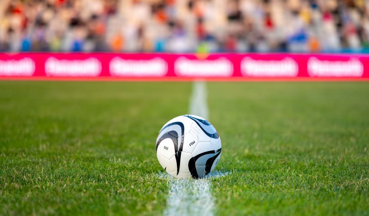 A soccer ball on the halfway line with a crowd in the background.