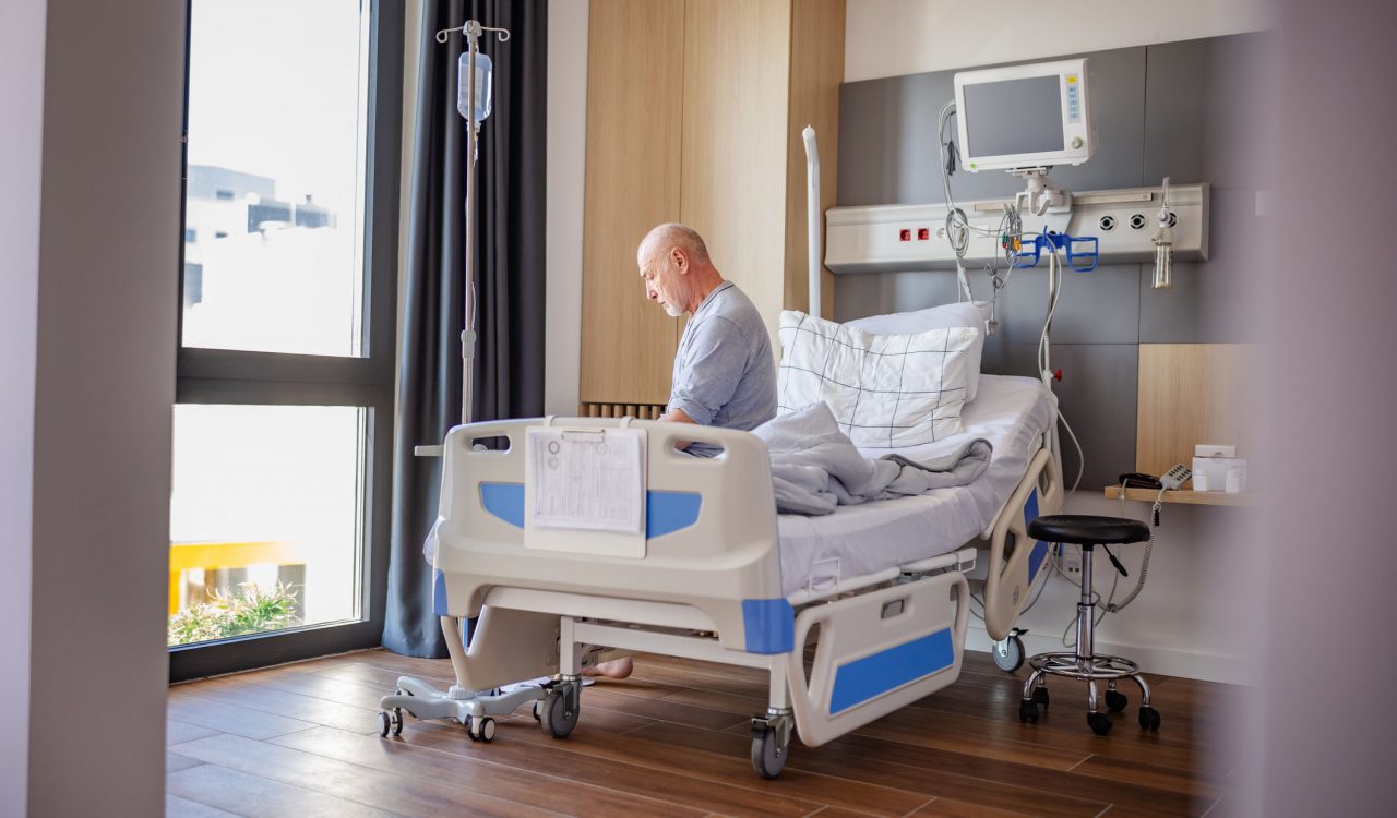 An older man sits on a hospital bed in a sunny hospital room.