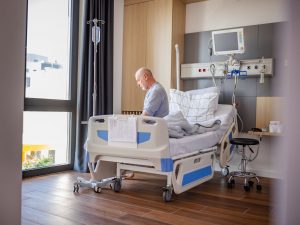An older man sits on a hospital bed in a sunny hospital room.