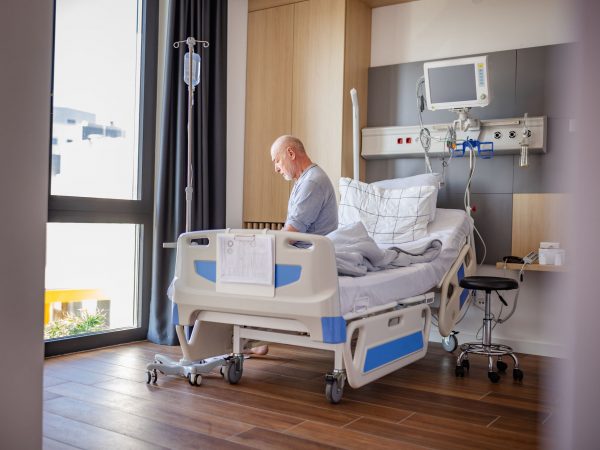 An older man sits on a hospital bed in a sunny hospital room.