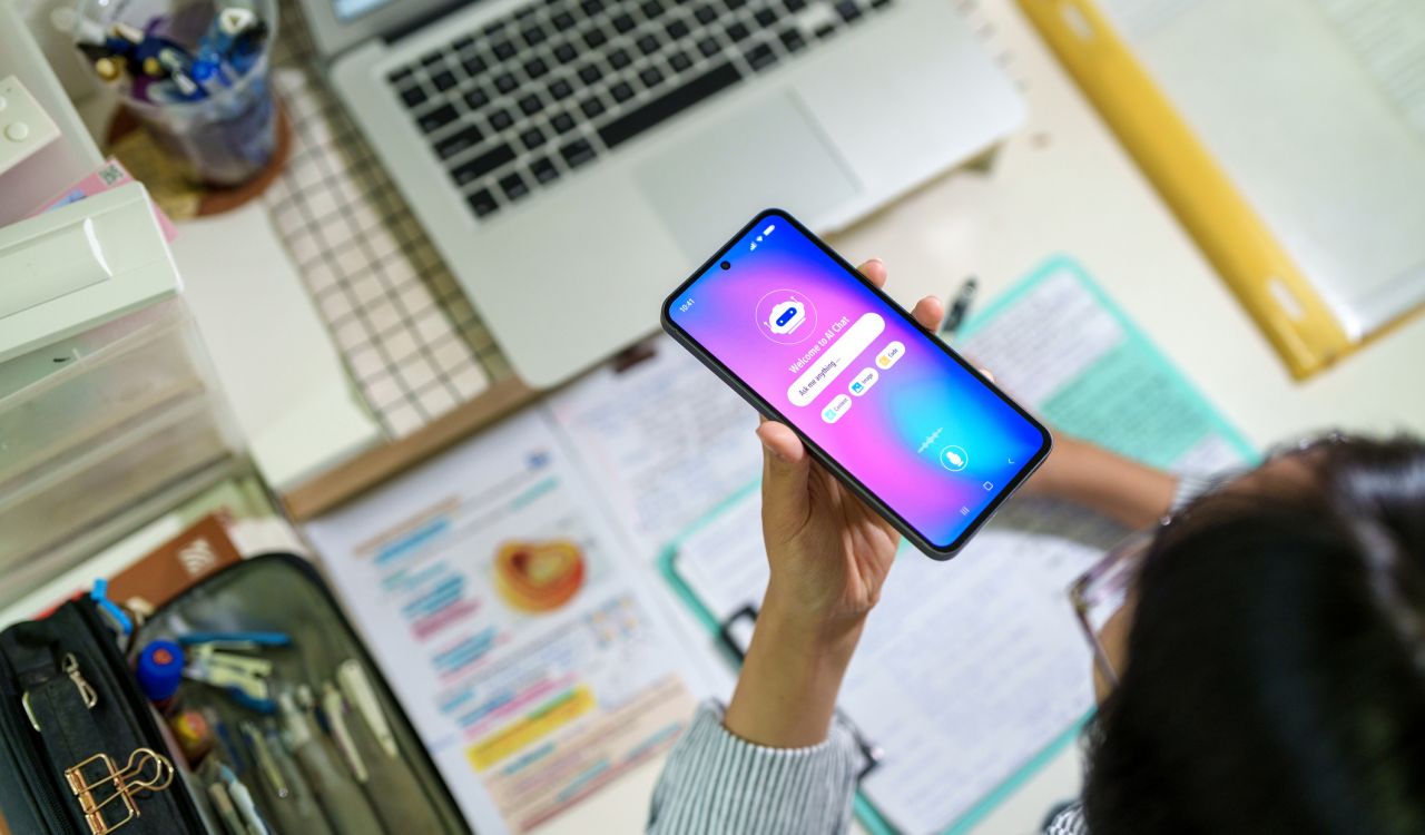 A teenage student interacts with an AI chatbot on a smartphone while studying at a desk with a laptop, notes and stationery. The scene highlights modern learning and technology integration.
