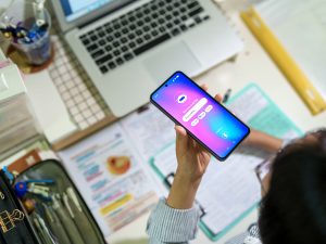 A teenage student interacts with an AI chatbot on a smartphone while studying at a desk with a laptop, notes and stationery. The scene highlights modern learning and technology integration.