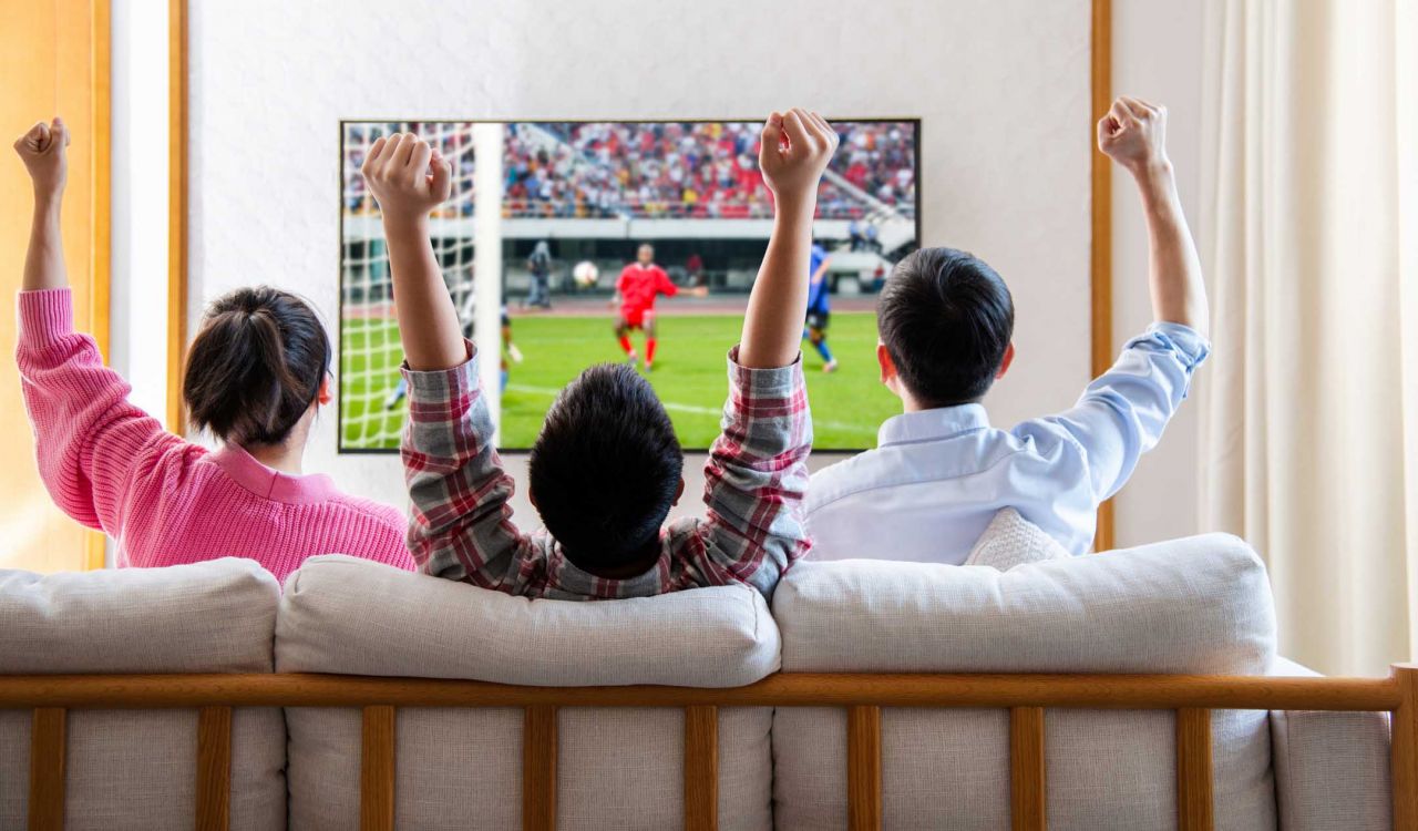 Three people sit on a sofa watching soccer on a TV.