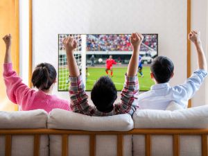 Three people sit on a sofa watching soccer on a TV.