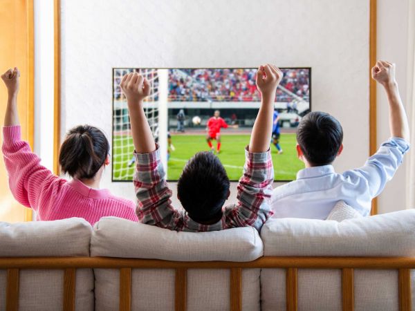 Three people sit on a sofa watching soccer on a TV.