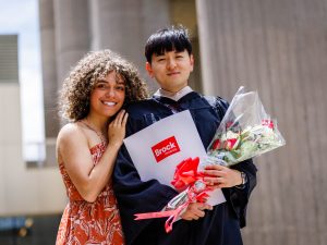 A new university graduate in an academic robe poses for a photo with loved one while holding flowers and a diploma.