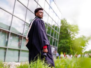 A young man in an academic robe stands in front of a glass building after a university graduation ceremony.