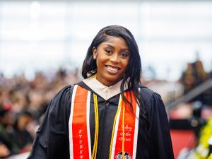 A young woman in academic robes pauses to smile for a photo after walking across the stage during a university graduation ceremony.