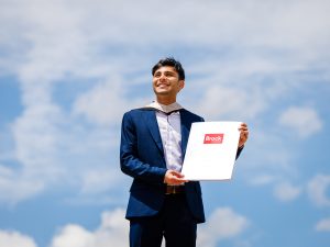 A young man in a suit poses with a diploma in front of a sky-themed backdrop after a university graduation ceremony.