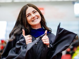 A young woman in an academic robe give two thumbs up and smiles at a graduation ceremony.