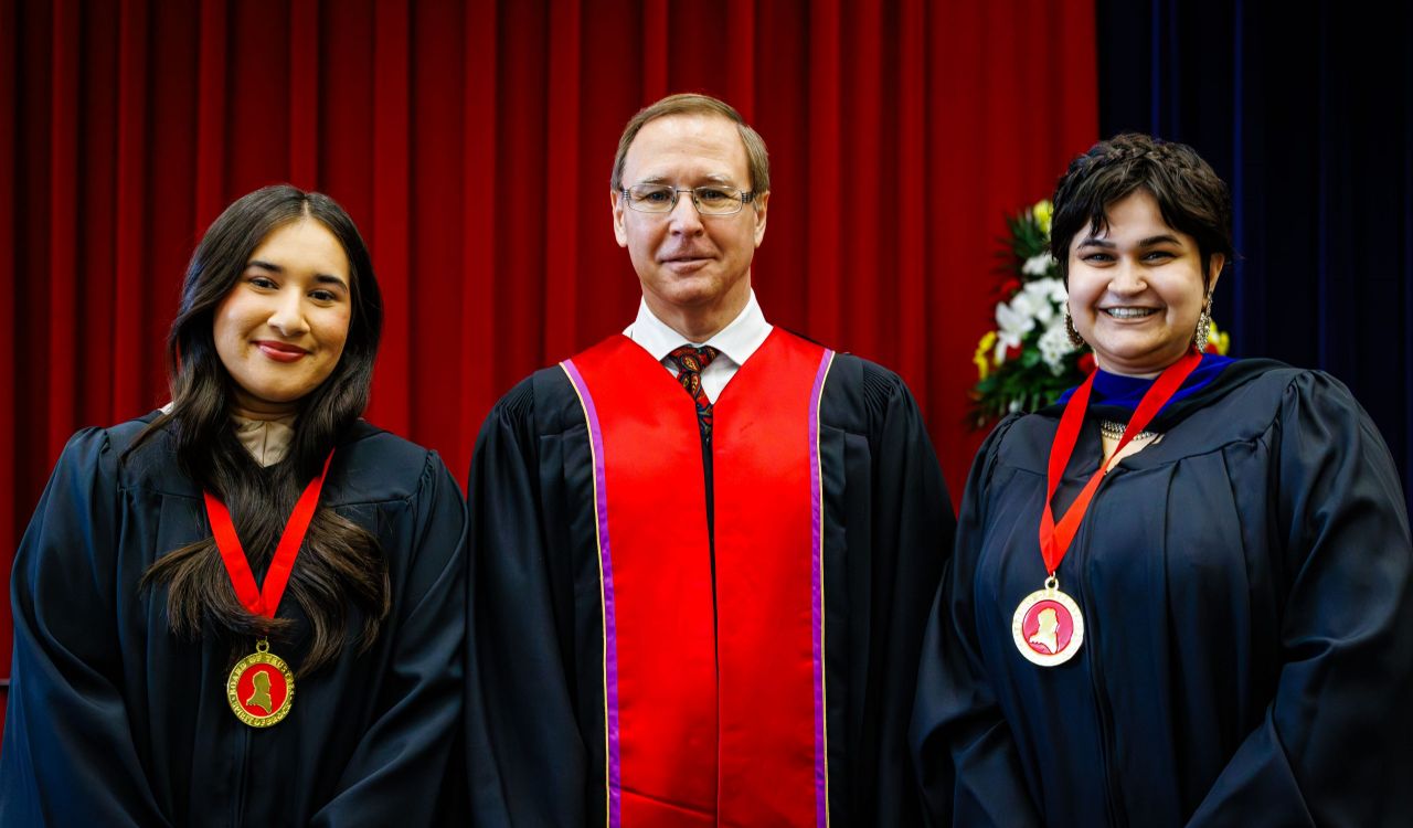 Three people in academic robes pose on stage for a photo during a university graduation ceremony.