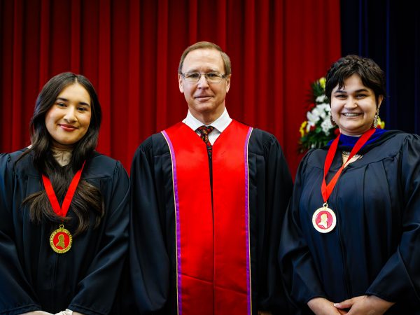 Three people in academic robes pose on stage for a photo during a university graduation ceremony.