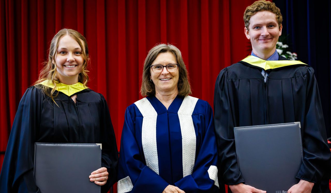 Three people in academic robes pose for a photo on stage during a university graduation ceremony.