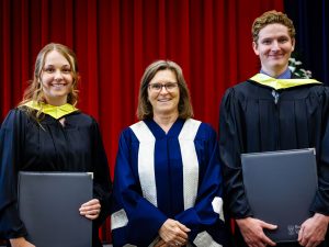 Three people in academic robes pose for a photo on stage during a university graduation ceremony.