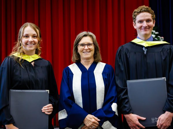 Three people in academic robes pose for a photo on stage during a university graduation ceremony.