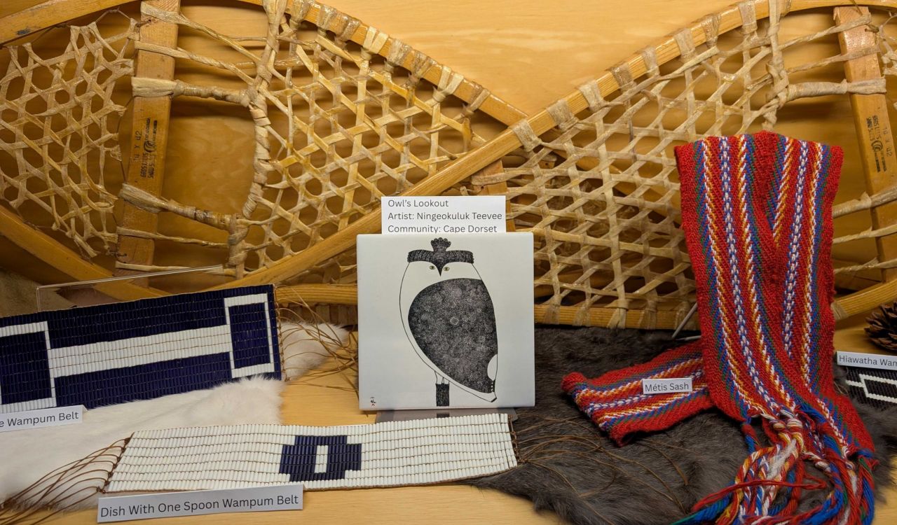 Two wooden snowshoes hang on a wall above a display of Indigenous cultural items, including beaded belts, a red-and-blue woven sash, a piece of art, and fur pelts.