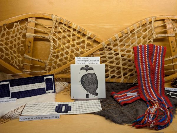 Two wooden snowshoes hang on a wall above a display of Indigenous cultural items, including beaded belts, a red-and-blue woven sash, a piece of art, and fur pelts.