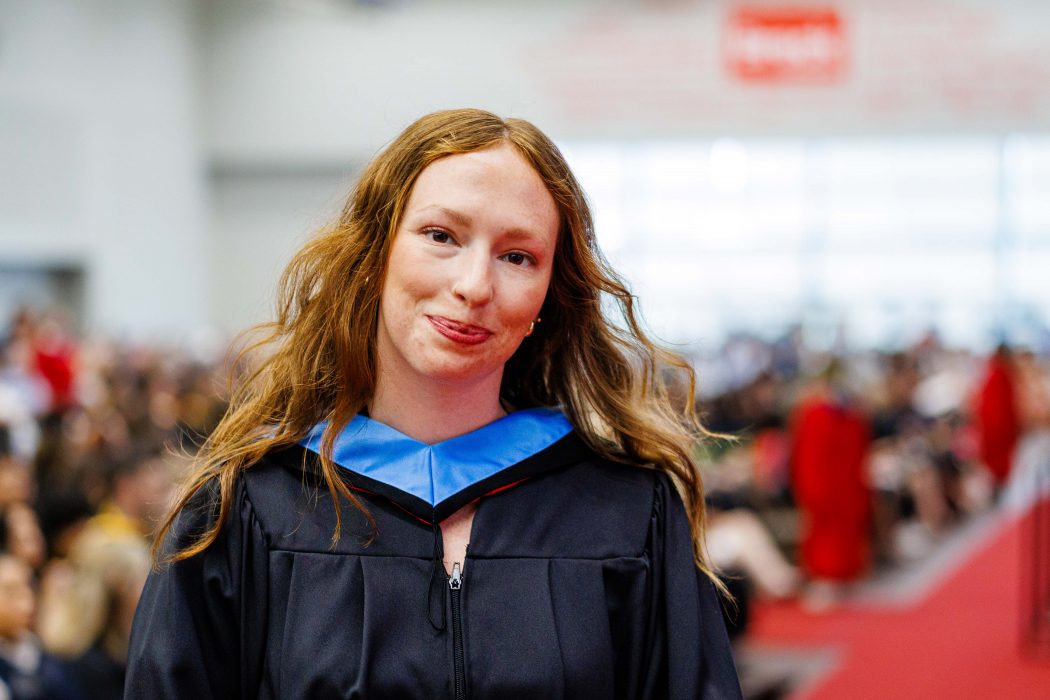 A young woman in academic gown smiles while standing in a university graduation ceremony.