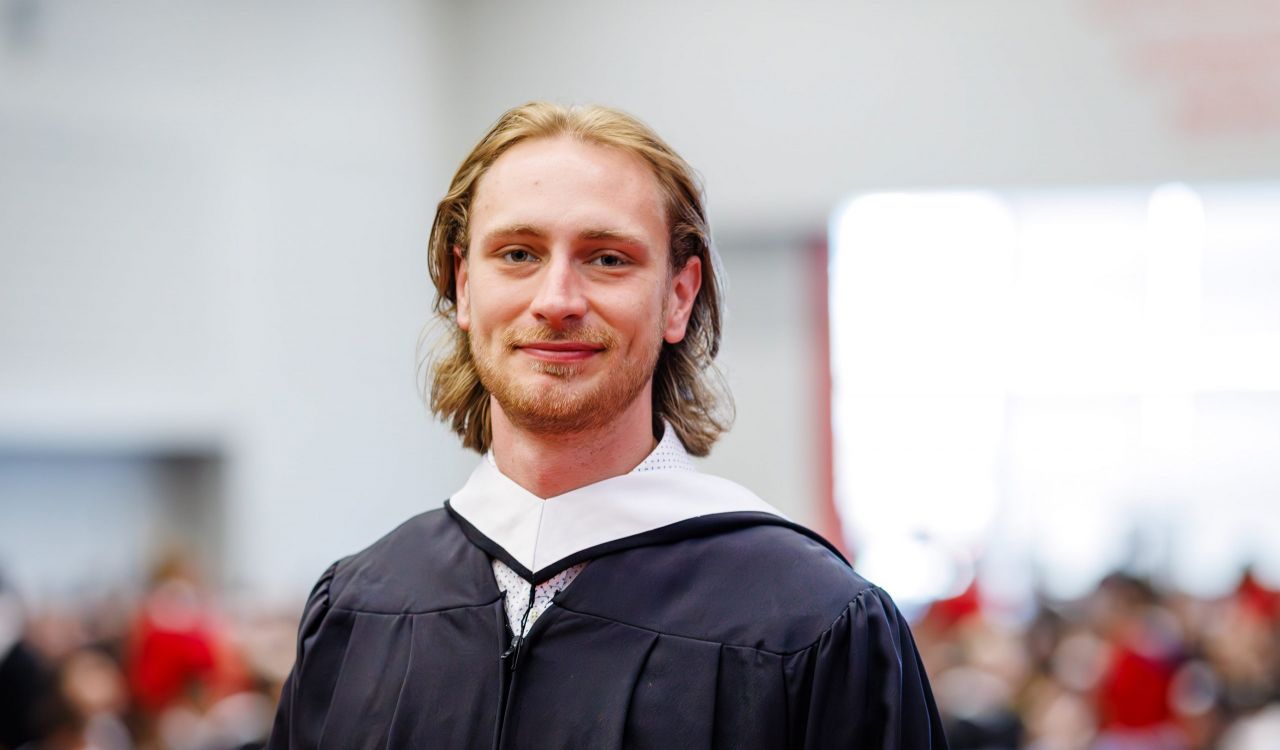 A young man in an academic robe smiles while standing in front of an audience at a university graduation ceremony.