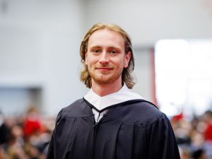 A young man in an academic robe smiles while standing in front of an audience at a university graduation ceremony.