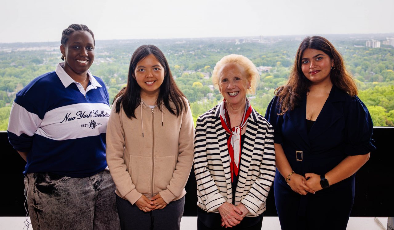 Four people stand side by side on a balcony, smiling at the camera, with a panoramic view of a leafy urban landscape behind.