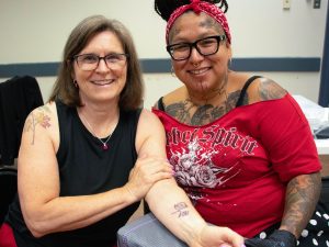 Two women sit side by side indoors. The woman on the left has her arm outstretched drawing your attention to a new tattoo on her forearm in the shape of a flower.