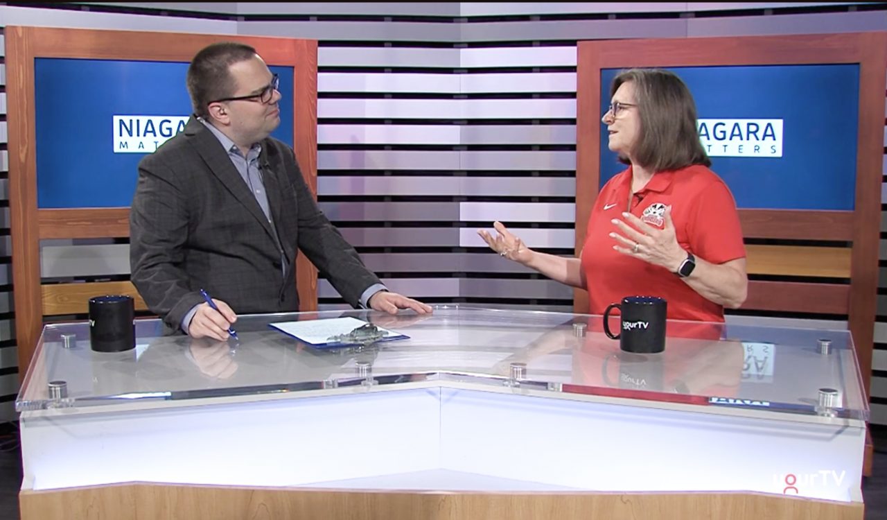 Two people stand at news desks speaking to one another.
