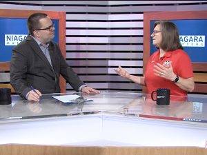 Two people stand at news desks speaking to one another.