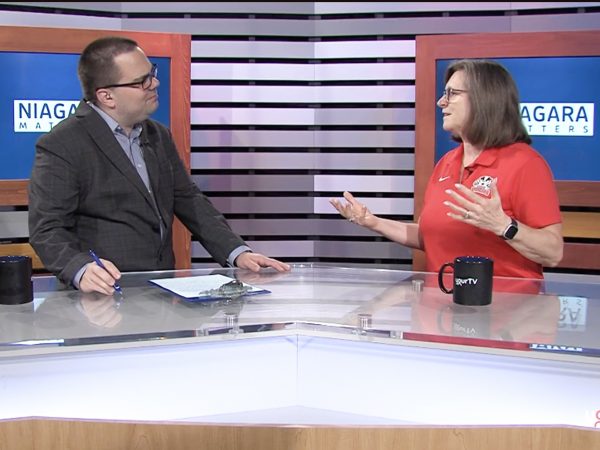 Two people stand at news desks speaking to one another.