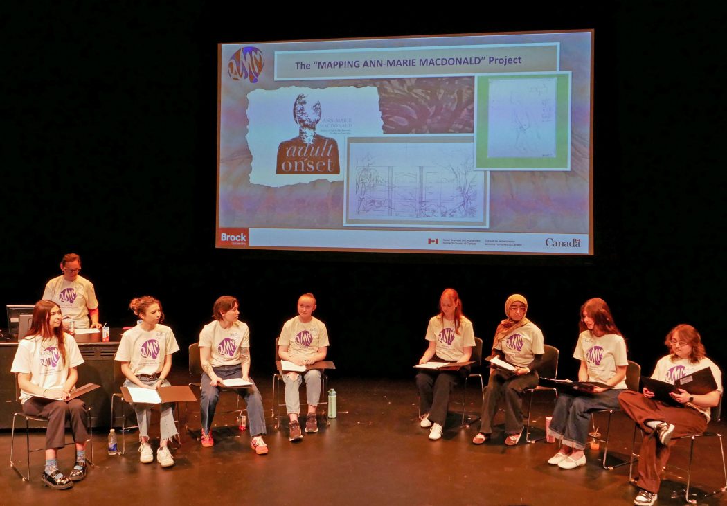 A group of people sit in chairs on a stage while a projector screen showcasing the Mapping Ann-Marie MacDonald Project shows a collage of illustrations.