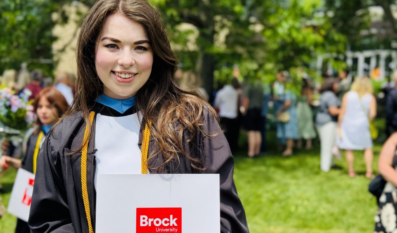 A young woman in an academic robe stands and smiles while holding a diploma outside a university graduation ceremony.