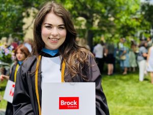 A young woman in an academic robe stands and smiles while holding a diploma outside a university graduation ceremony.