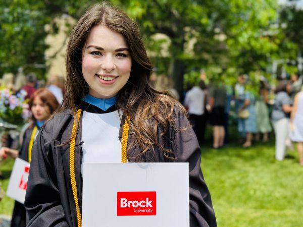 A young woman in an academic robe stands and smiles while holding a diploma outside a university graduation ceremony.